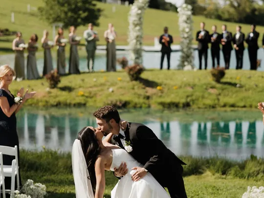 A groom dips his bride on the pond island with the wedding party lined along the water at this New Market farm wedding venue